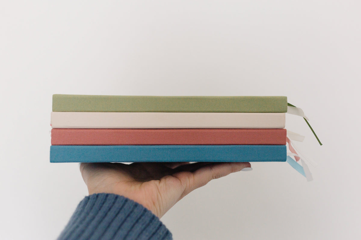Hand holding three linen notebooks with tied ribbon, green, terracotta, and teal covers