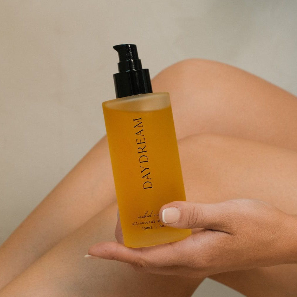 Amber body oil bottle with black pump held in hand, bare leg visible.