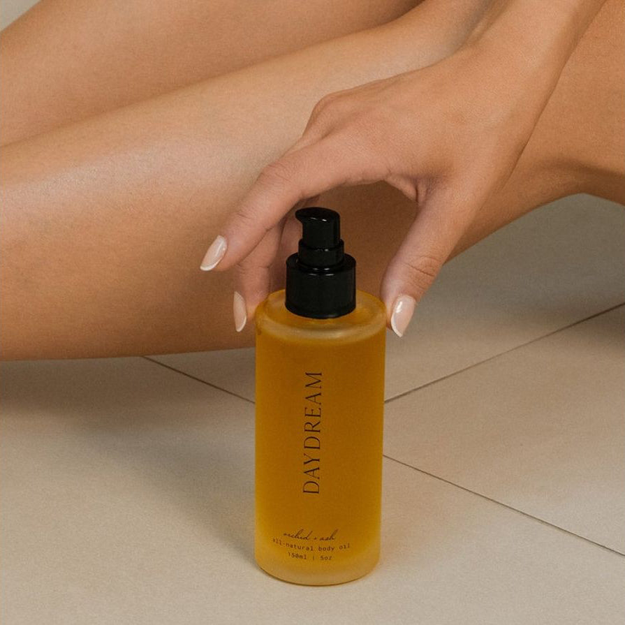 Amber body oil bottle with black pump held against bare skin for application.