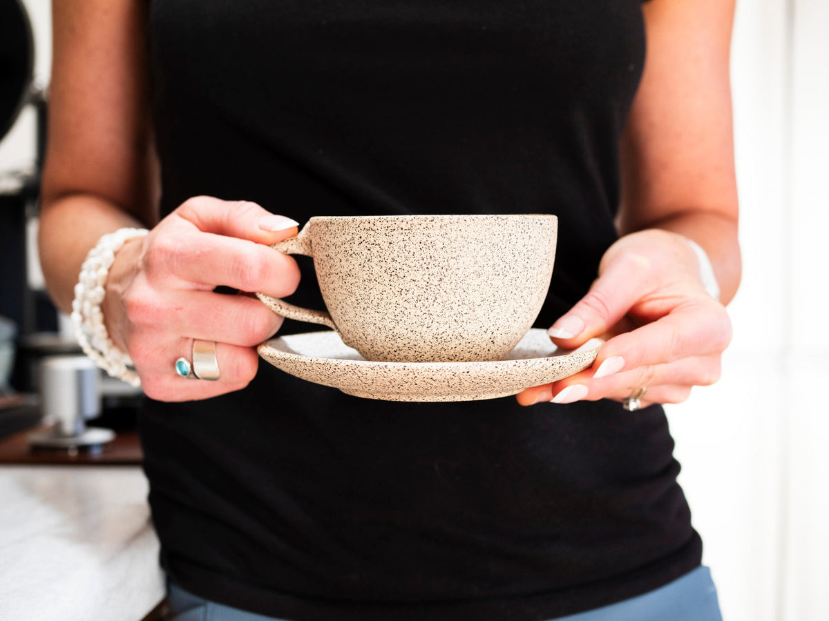 Speckled stoneware latte cup with handle on saucer, held by person