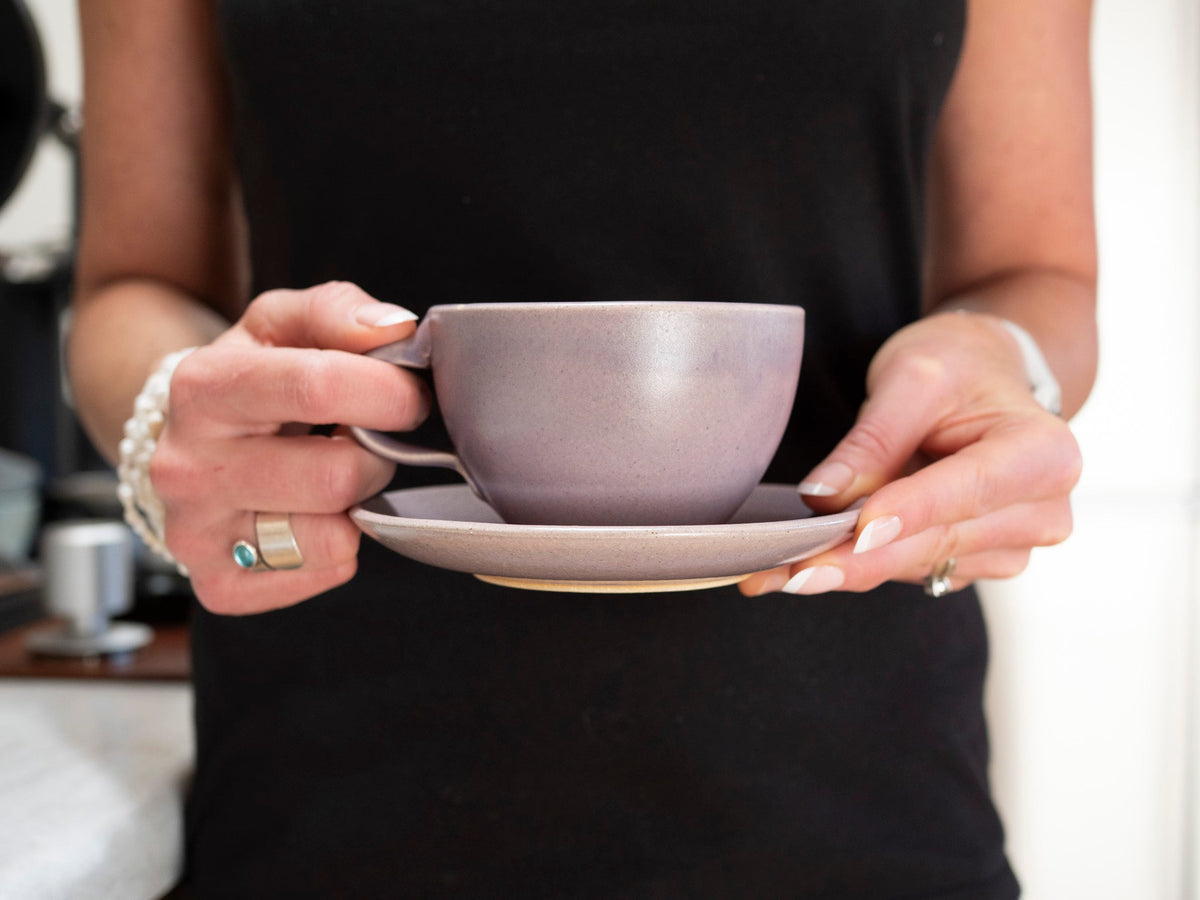 Purple stoneware cup with handle on matching saucer, held in hands.