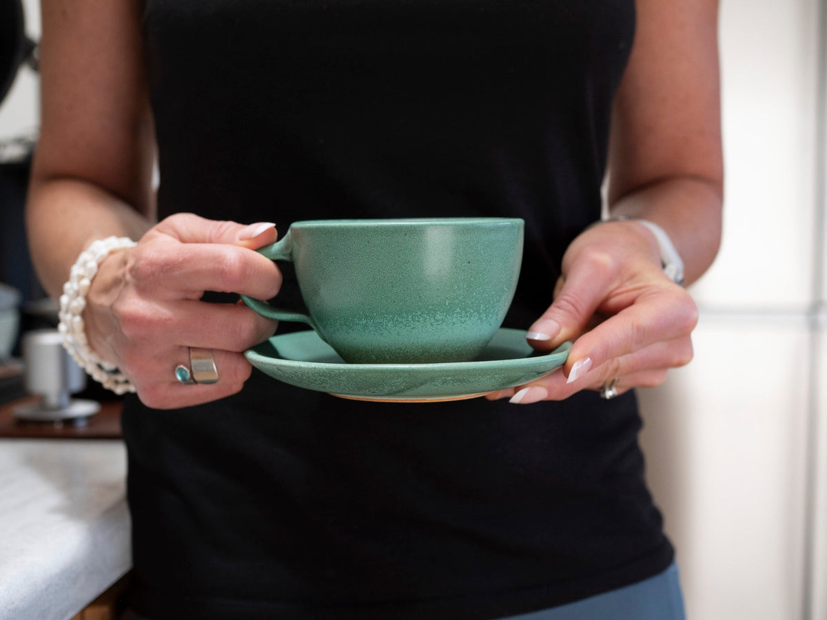 Hands holding a green stoneware latte cup with handle and matching saucer.