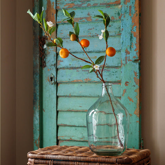 Tangerine branch stems with orange and yellow fruit, green leaves in clear glass bottle.