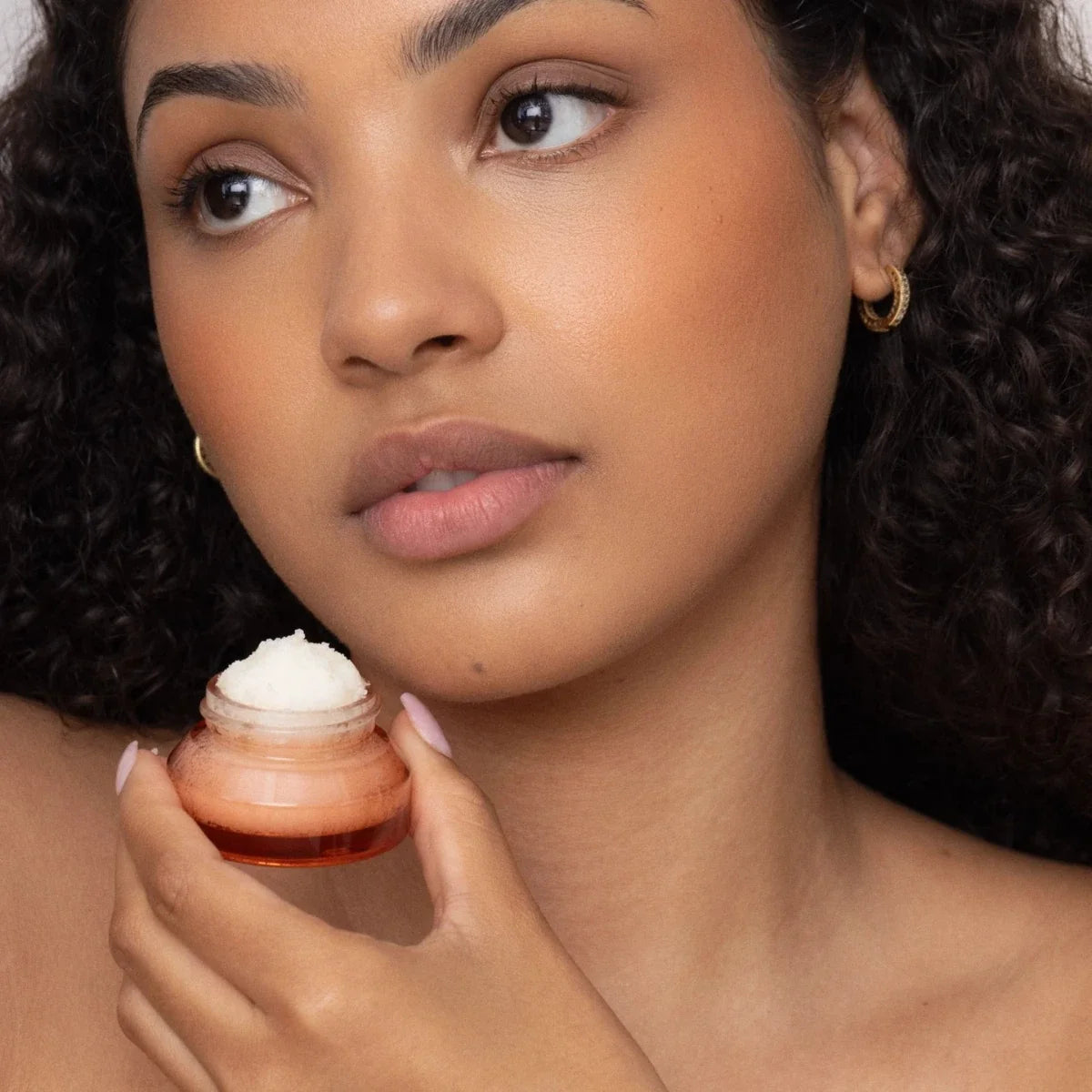 Lip scrub jar held near lips, showing white exfoliating scrub