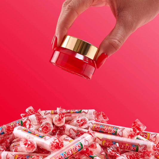 Red lip scrub jar with gold lid held above a pile of Smarties candies