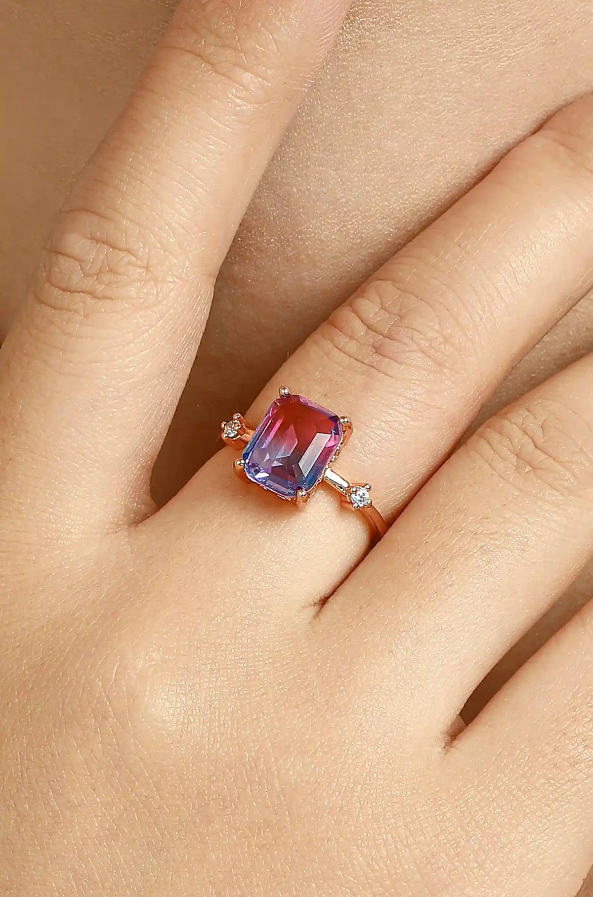 Rose gold ring with a rectangular pink and violet sapphire and small clear side stones, worn on a hand