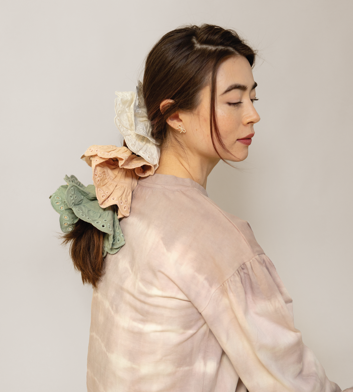 Eyelet scrunchies in cream, peach, and sage green worn together in a ponytail.