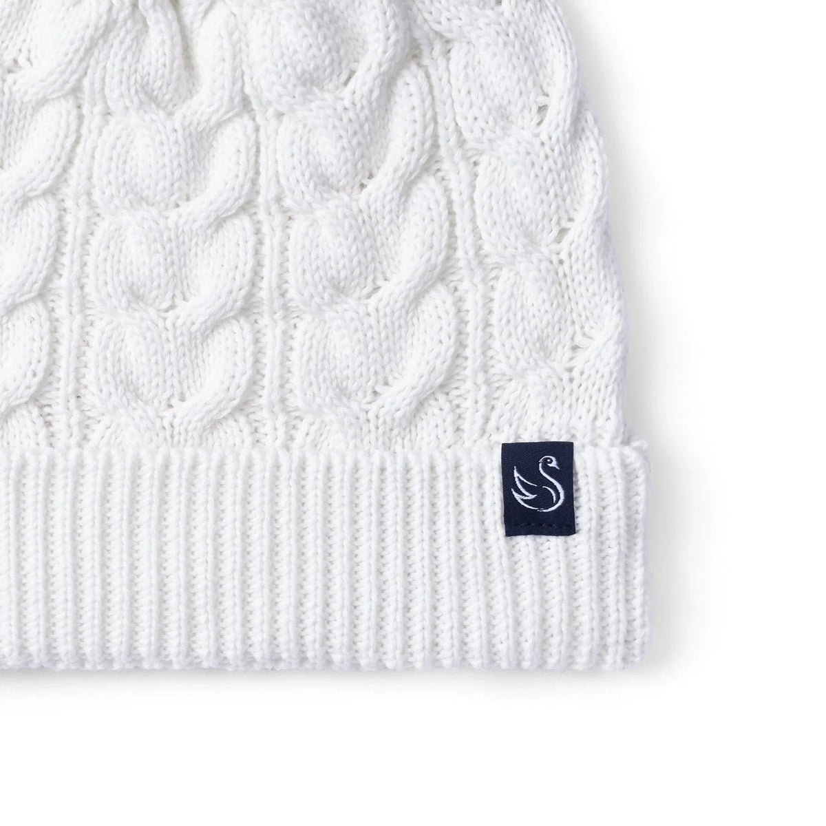 White cable knit beanie with pom-pom and navy logo patch on ribbed cuff.