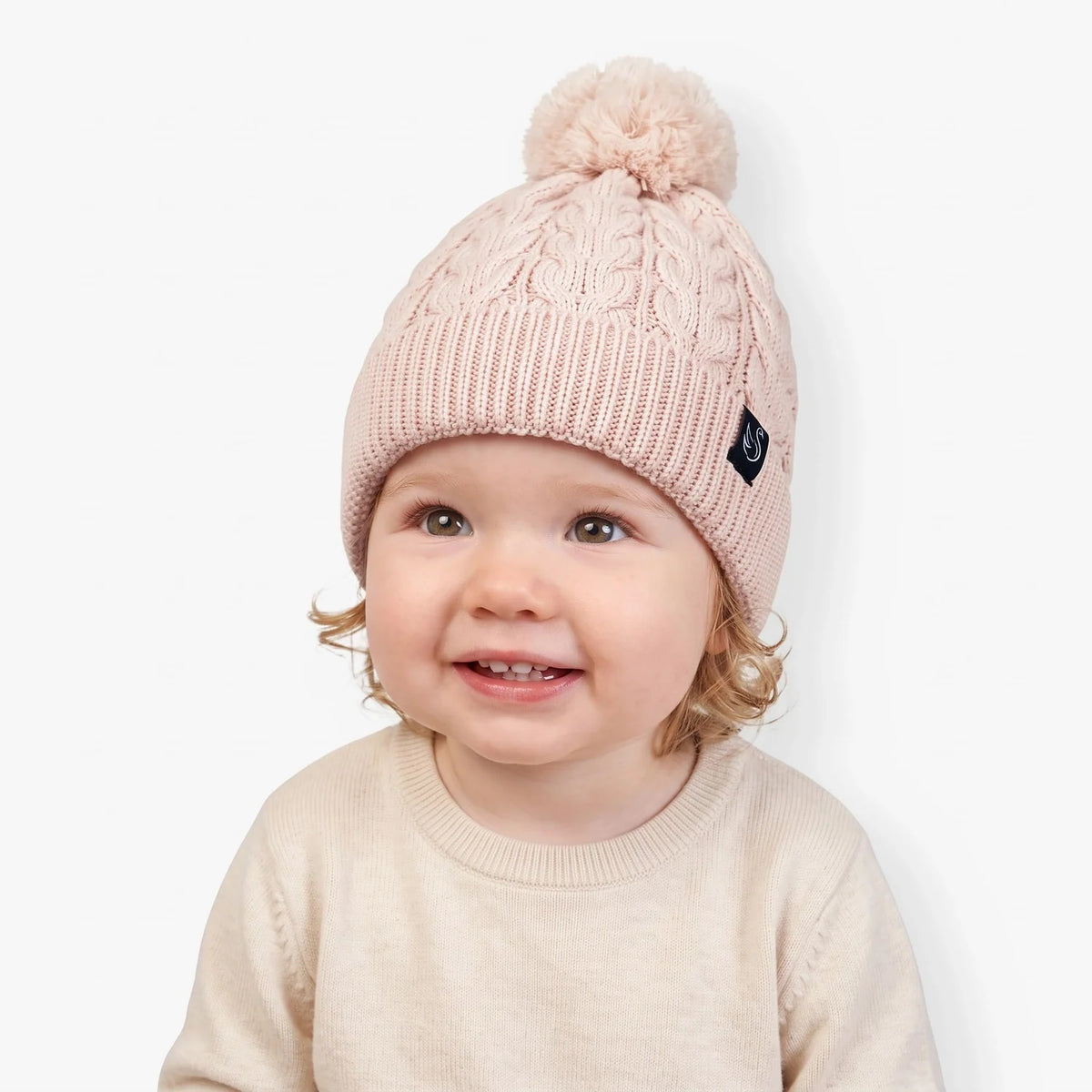 Beige cable-knit beanie with pom-pom and dark logo patch worn by a baby