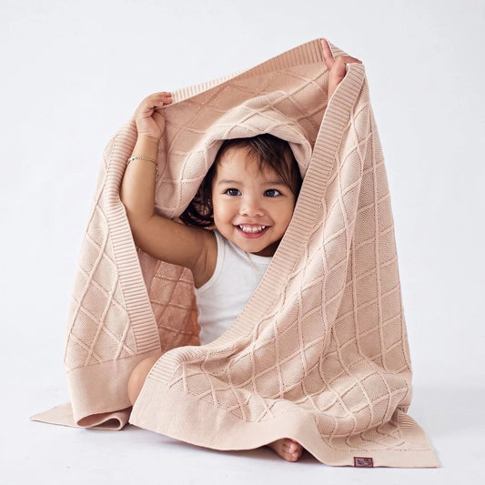 Beige knit baby blanket with diamond pattern, pulled over a smiling baby