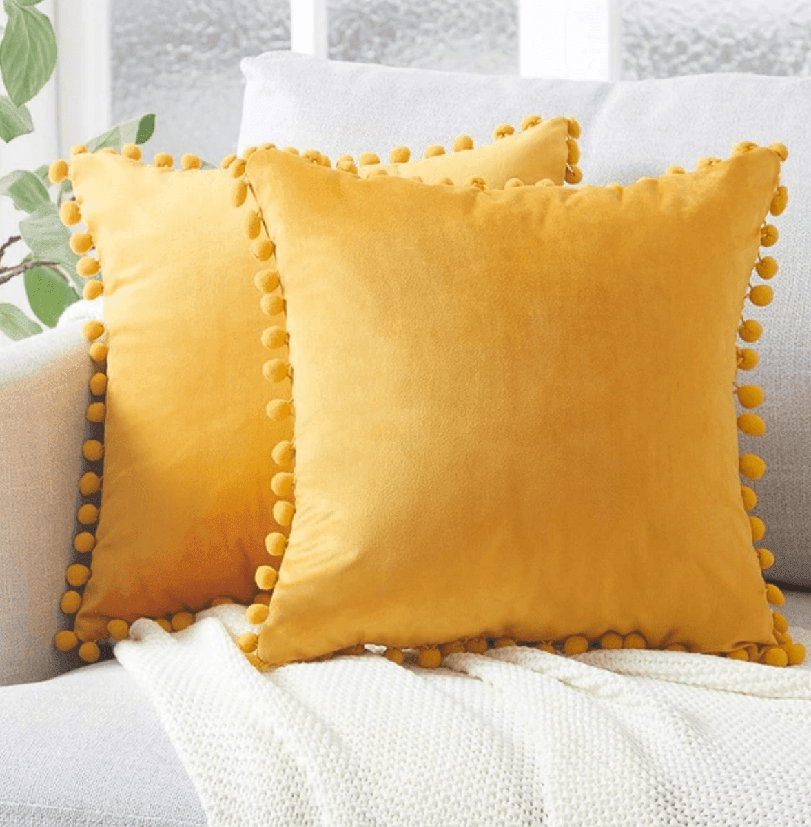 Mustard yellow plush throw pillow with pom-pom trim on a white bed