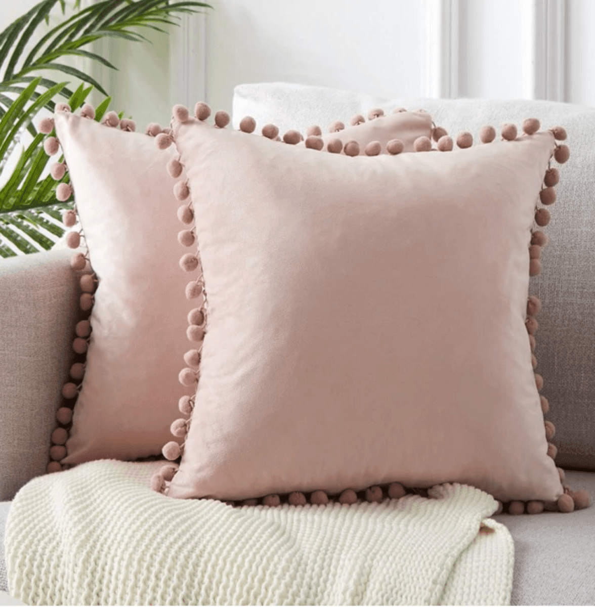 Cream plush throw pillow with pom-pom trim and a knit blanket nearby