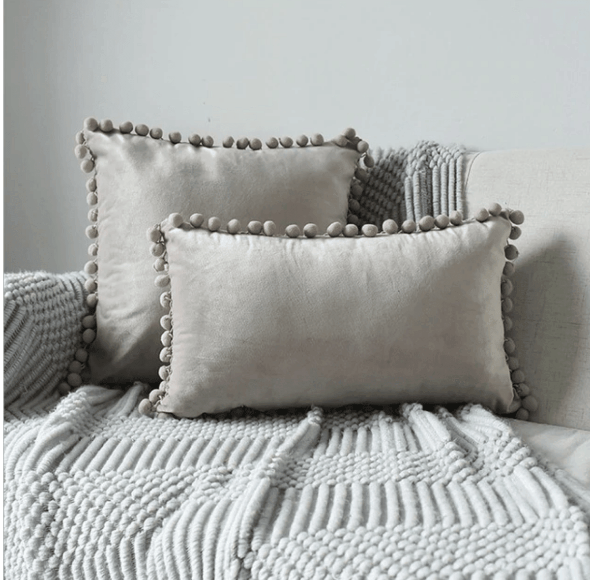 Light gray plush throw pillow with pom-pom trim on a textured white blanket
