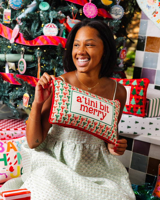 Mini needlepoint pillow held up: 'a tiny bit merry' red lettering with green border