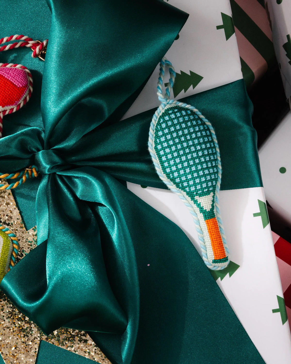 Small needlepoint tennis racket ornament on gift wrap, teal with orange handle.