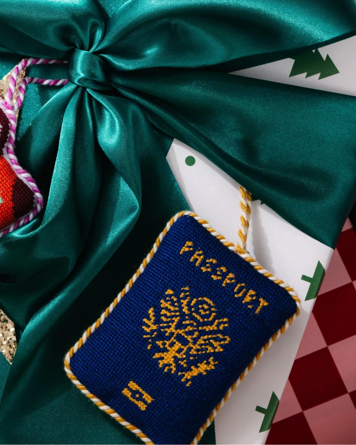 Blue needlepoint passport icon ornament on a wrapped gift, tied with gold-trim rope.