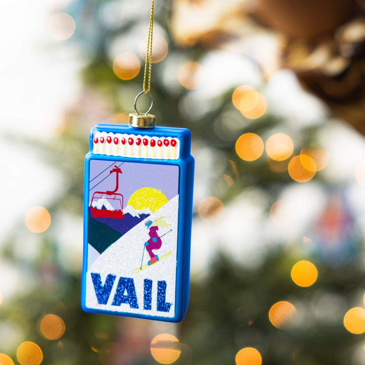 Blue matchbook-style ornament with VAIL logo, skier artwork, hanging on gold cord.