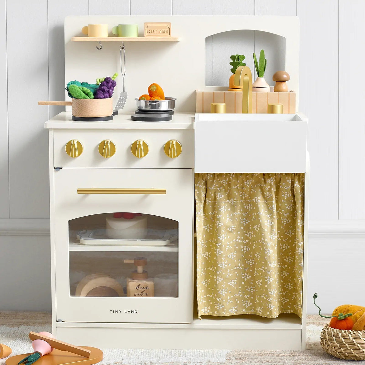 White wooden play kitchen with gold knobs, oven, sink, stovetop, and patterned storage curtain