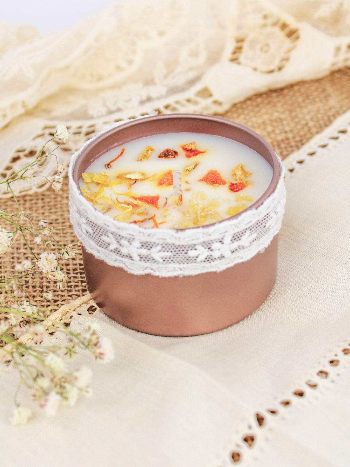 Rose-pink soy wax candle tin with open lid; white lace trim and dried orange botanicals