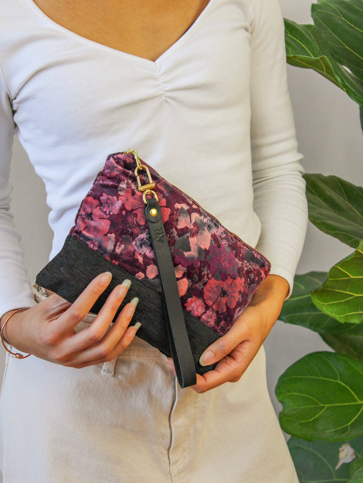 Velvet wristlet held at side, purple floral front, black accent panel, gold zipper pull.