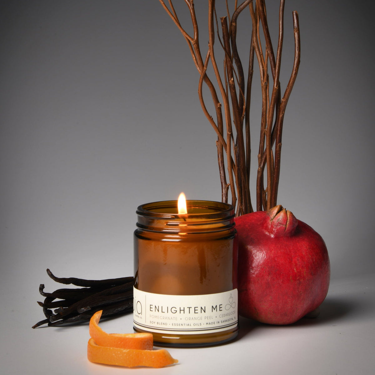 Soy blend candle in amber glass jar, flame lit, with pomegranate and orange peel decor