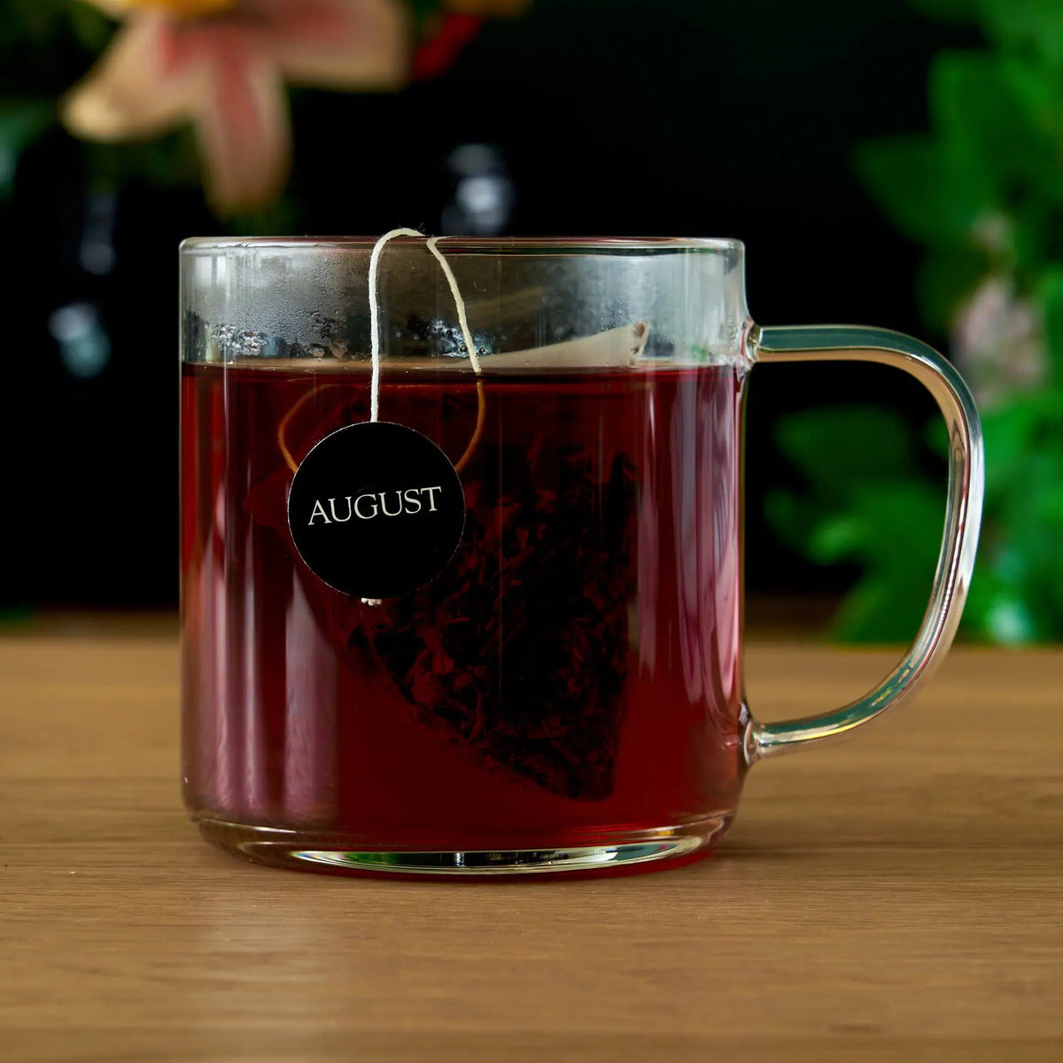 Brewed dark tea in a clear glass mug with a tea bag and August tag.