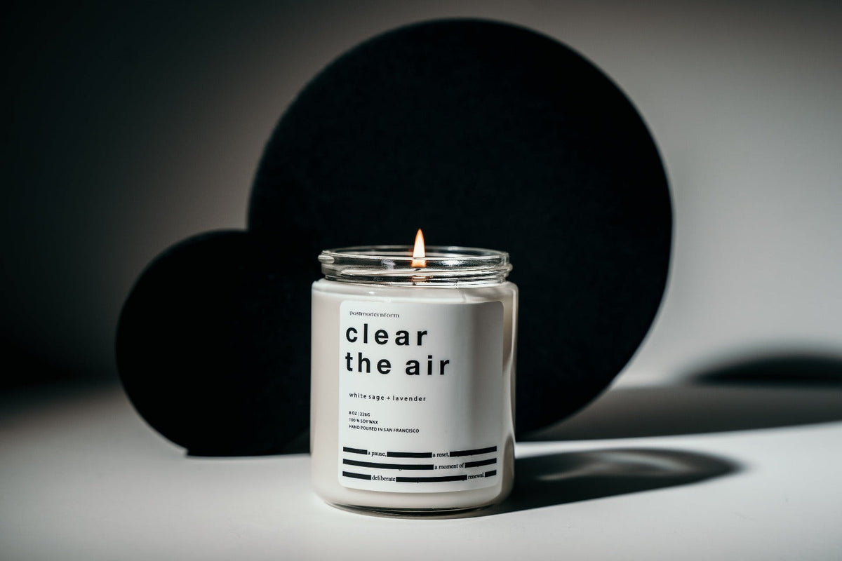 Small white candle jar with black lid, lit flame above clear the air label