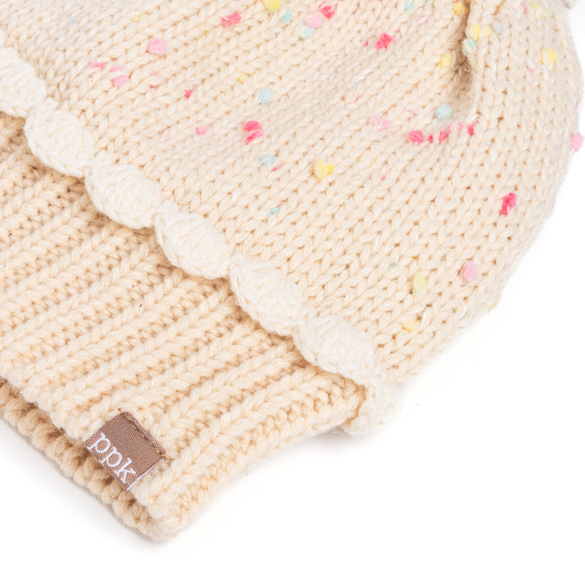 Close-up of pale knit beanie showing speckled yarn, scalloped trim, ribbed cuff, and label