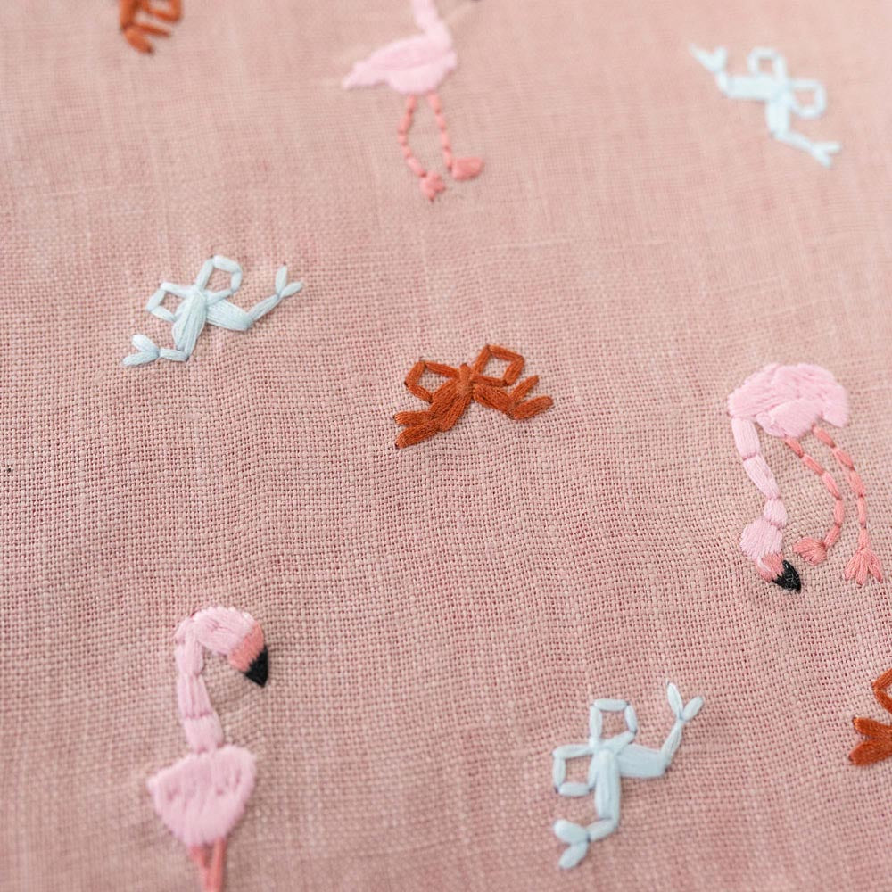 Beige stocking fabric close-up with embroidered pink, blue, and orange flamingos and bows