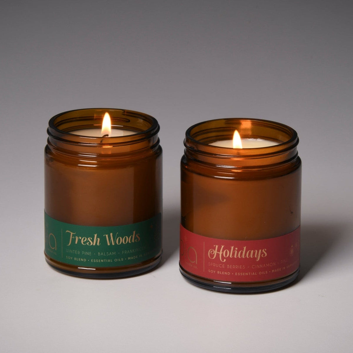 Close-up of two amber glass soy candles: green Fresh Woods label and red Holidays label, unlit
