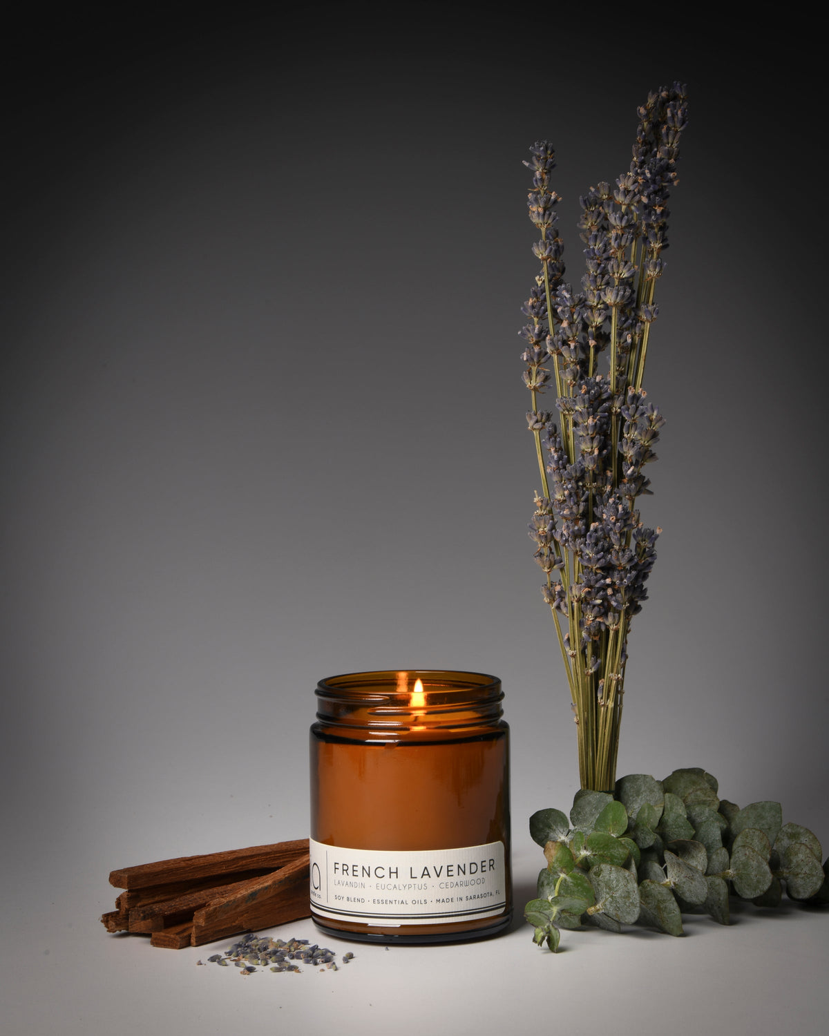 Amber soy candle jar with lit wick beside dried botanical stems and stones