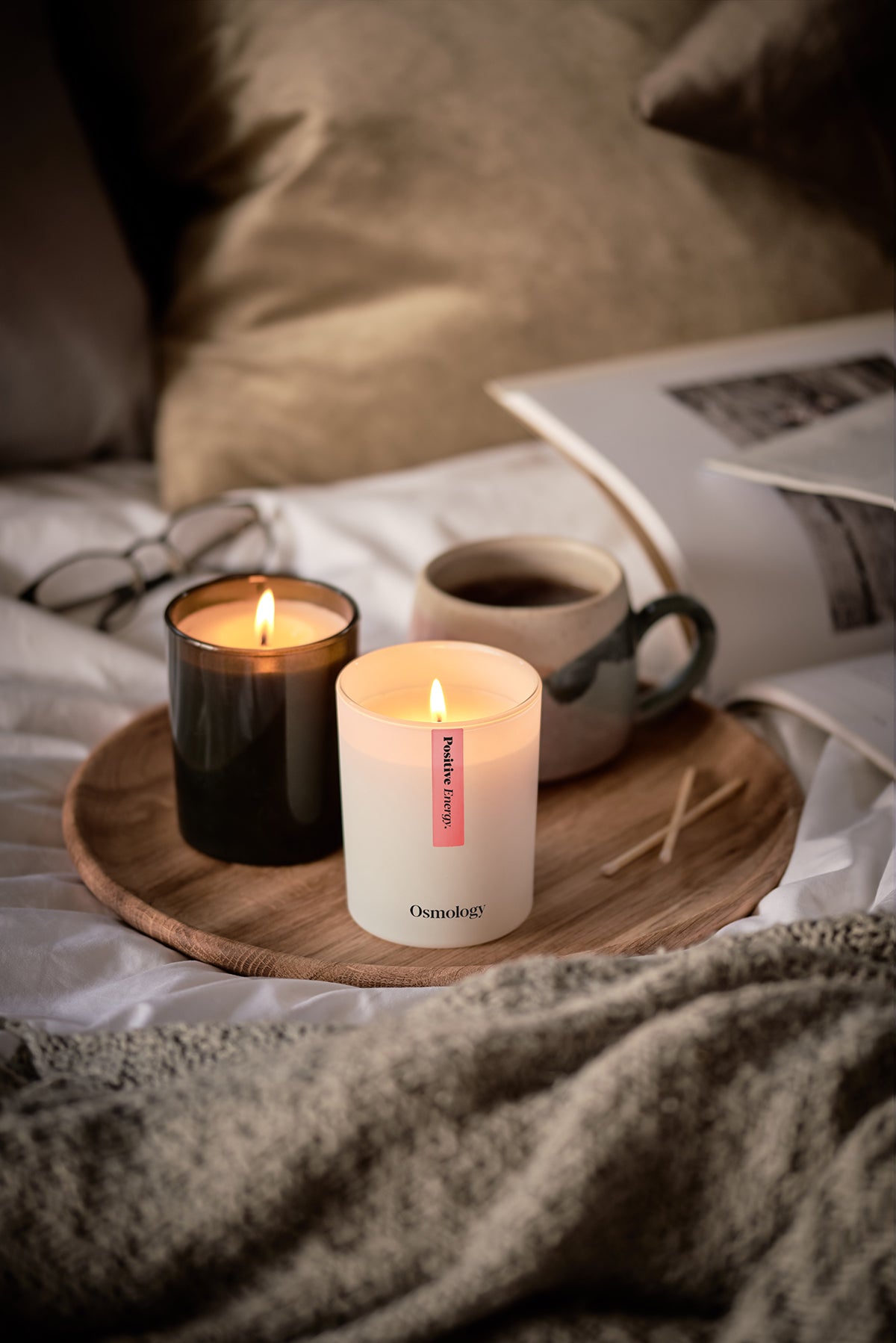 Two scented candles on wooden tray with coffee mugs, wicks lit (Osmology)