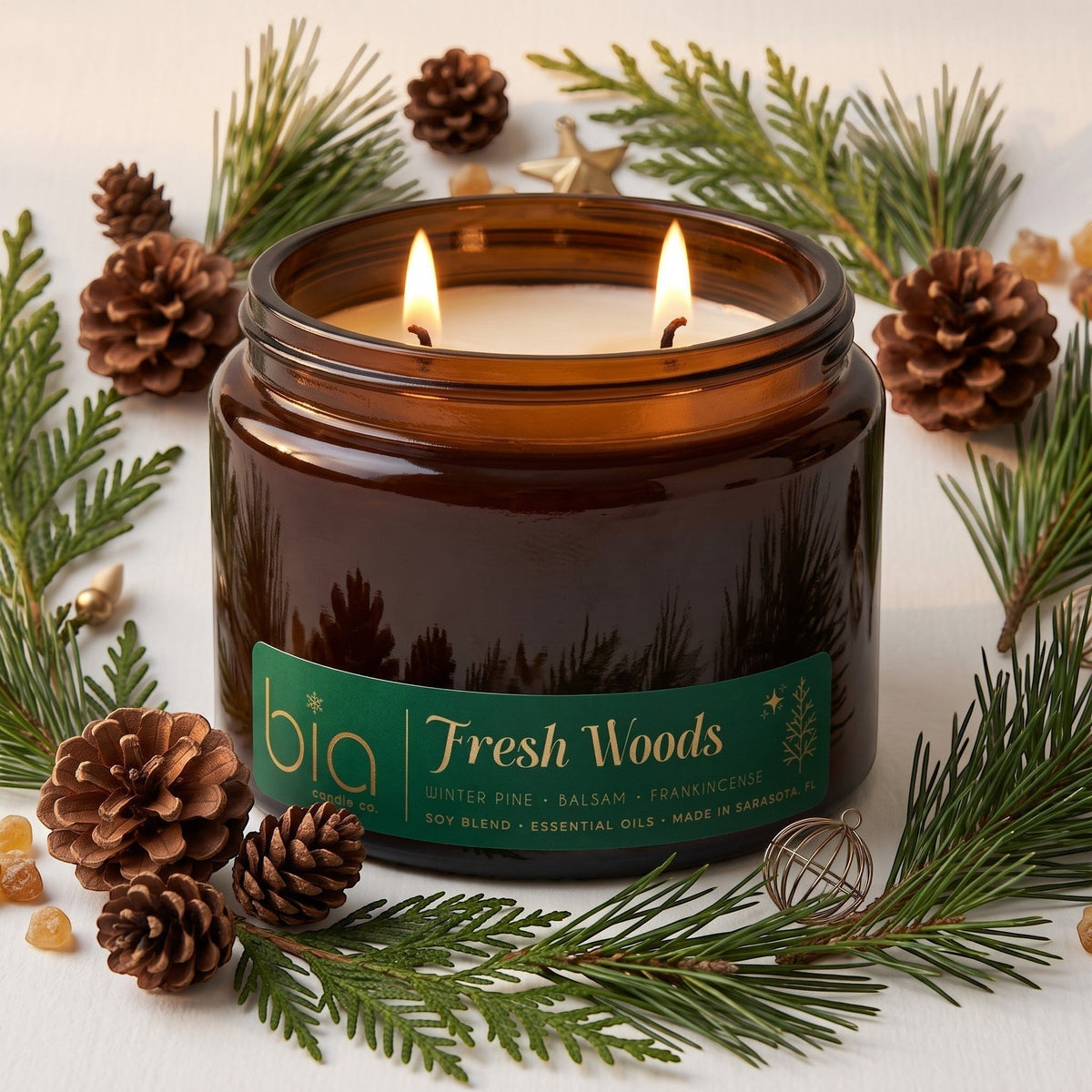 Amber glass candle jar with green label and lit flames, with pine branches and pinecones.
