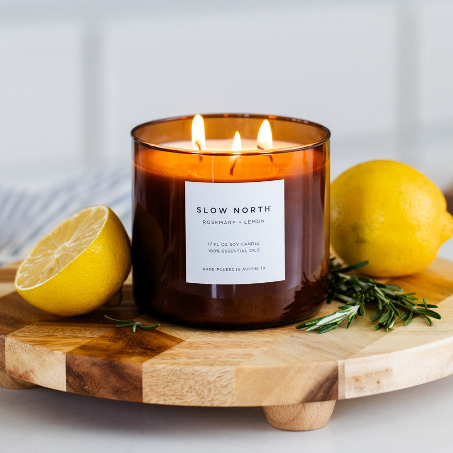Amber glass three-wick candle with lit wicks, rosemary, and lemon on wooden tray