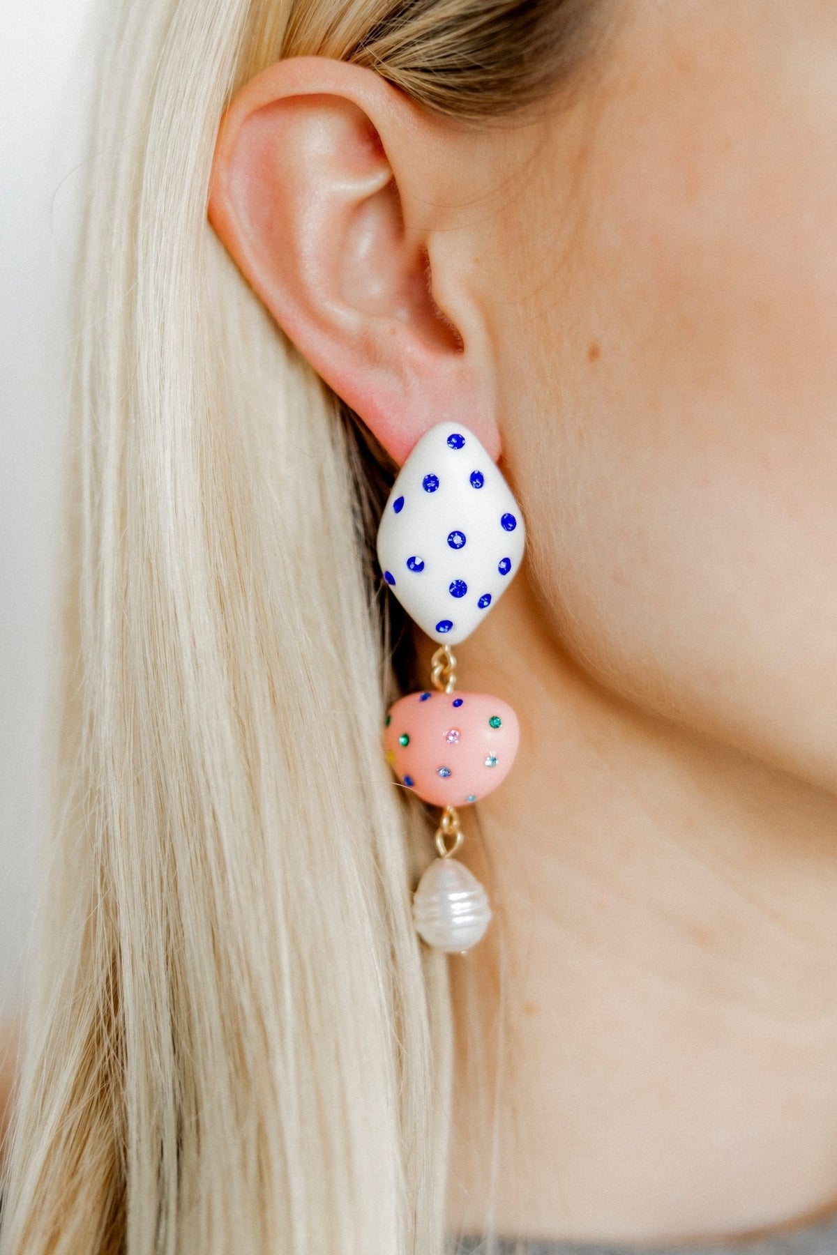 Gold-tone dangling earrings on ear: blue polka-dot teardrop, pink heart, pearl charm