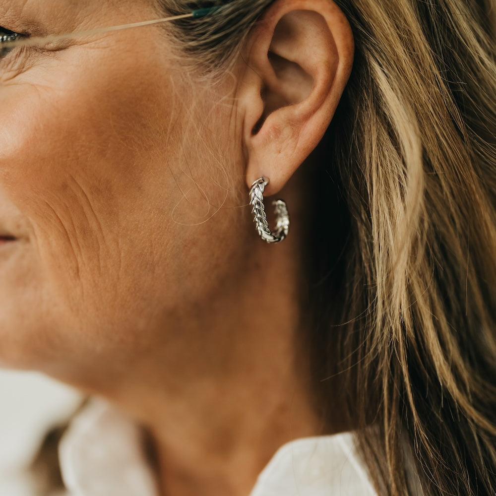 Silver braided oval hoop earrings on ear, twisted chain texture.