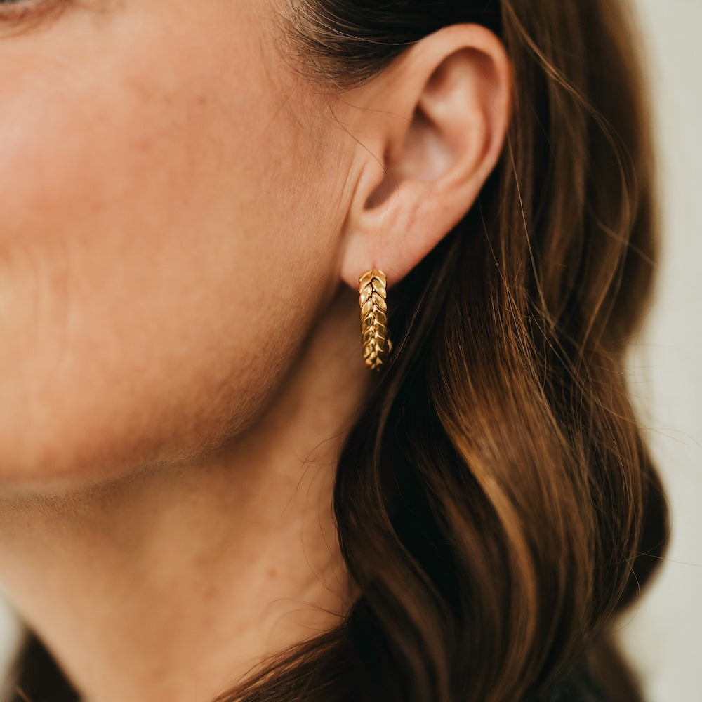 Gold braided oval hoop earrings on ear, twisted chain texture.