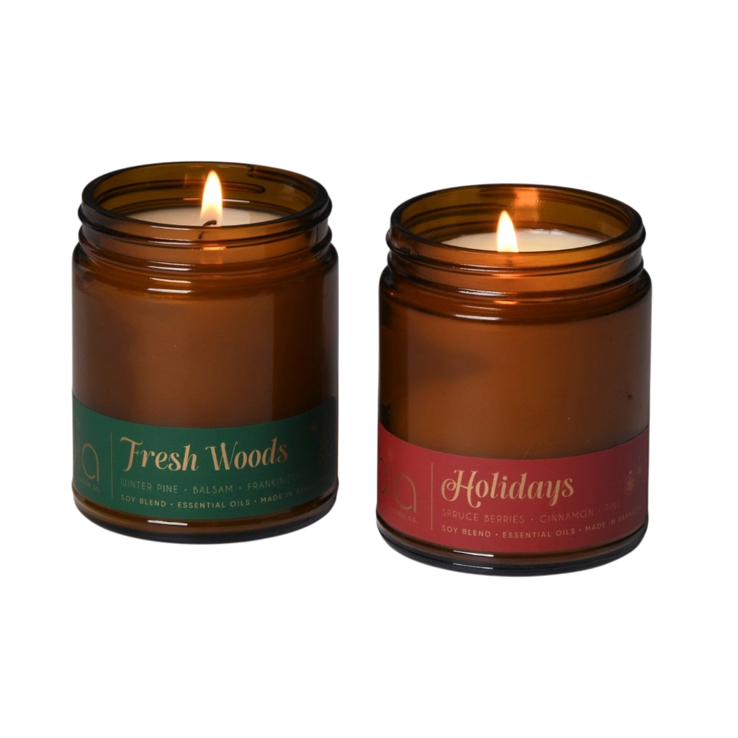Pair of amber glass soy candles with green Fresh Woods and red Holidays labels, both lit
