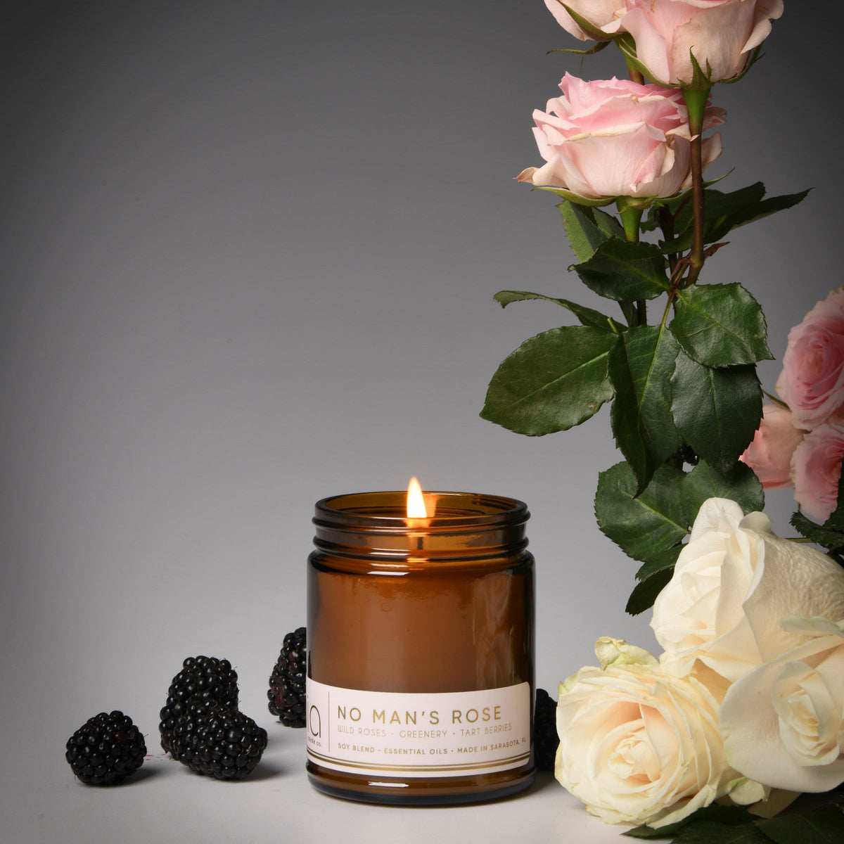Amber glass soy candle jar with lit wick and white label, dark leaf top edge visible.