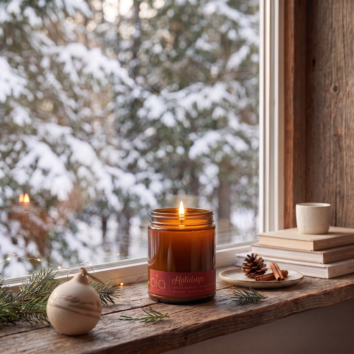 Lit amber candle jar on a windowsill, with pine branches and greenery nearby.