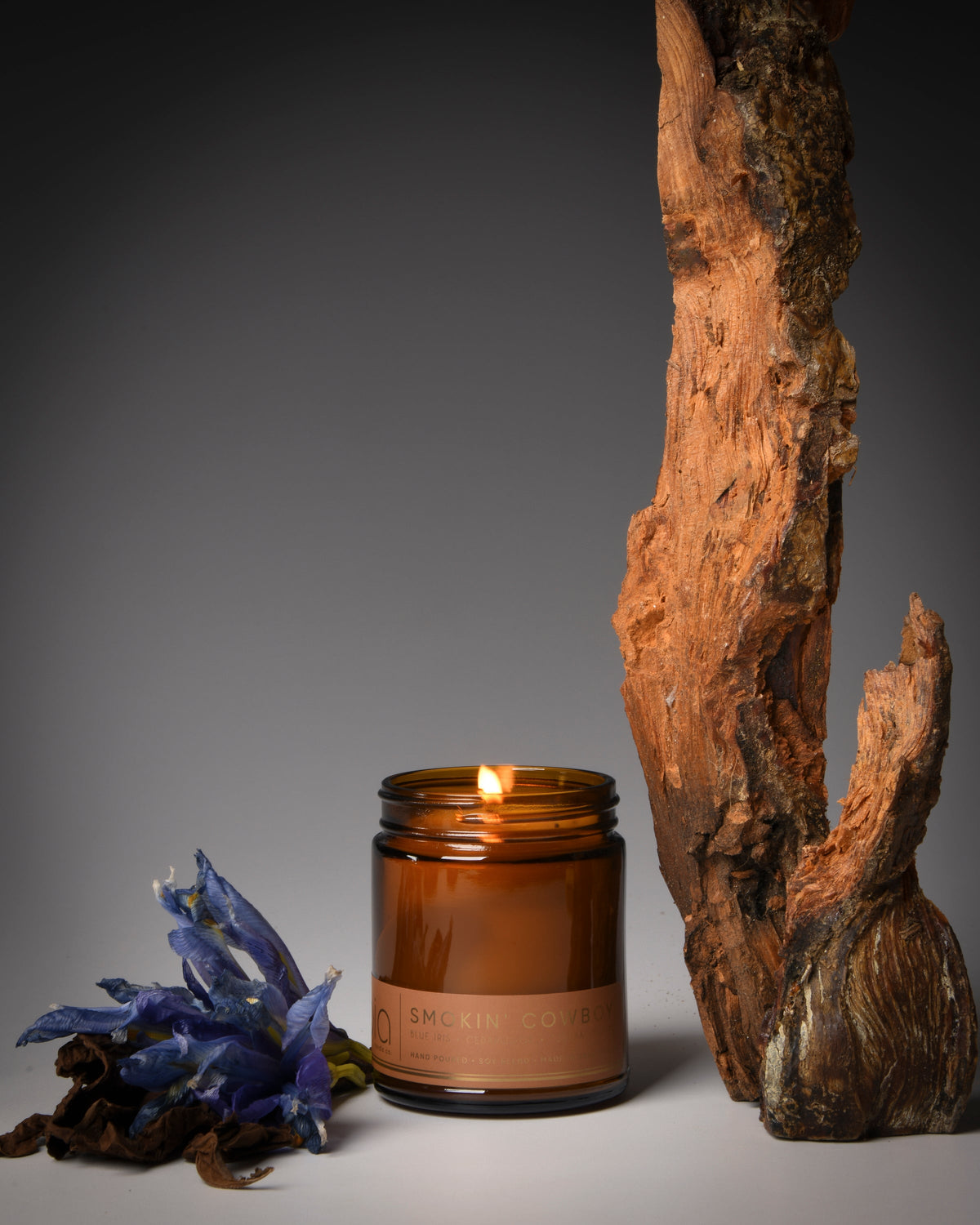 Amber glass candle with burning wick beside driftwood and blue flowers