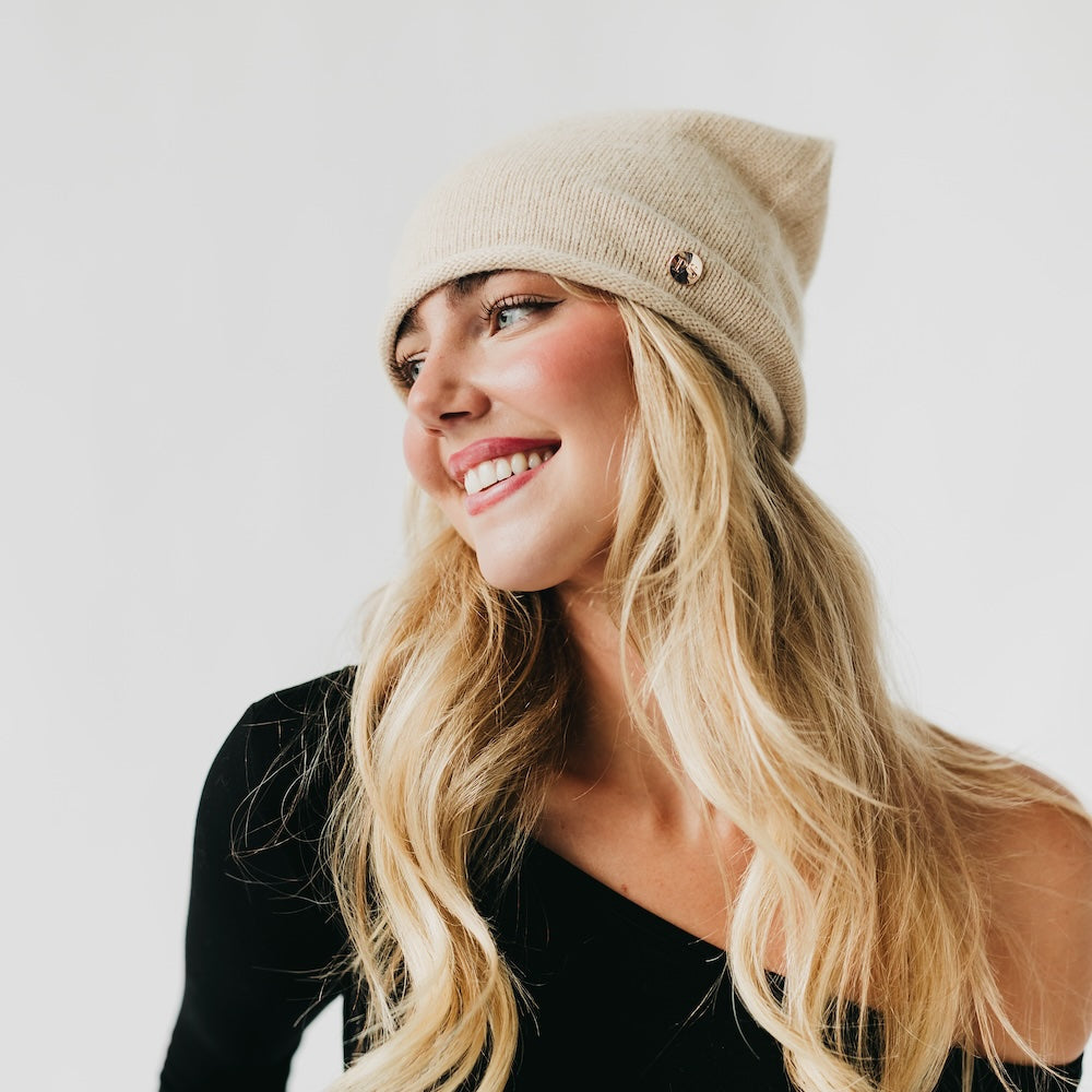 Beige cashmere beanie with rolled edge cuff and small front pin detail.