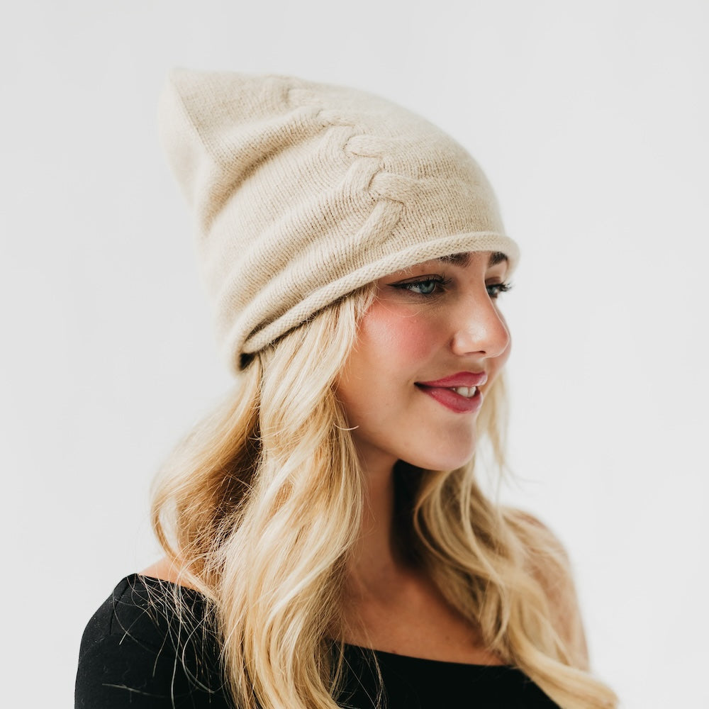 Beige cashmere beanie with rolled cuff and small front pin, angled close-up.