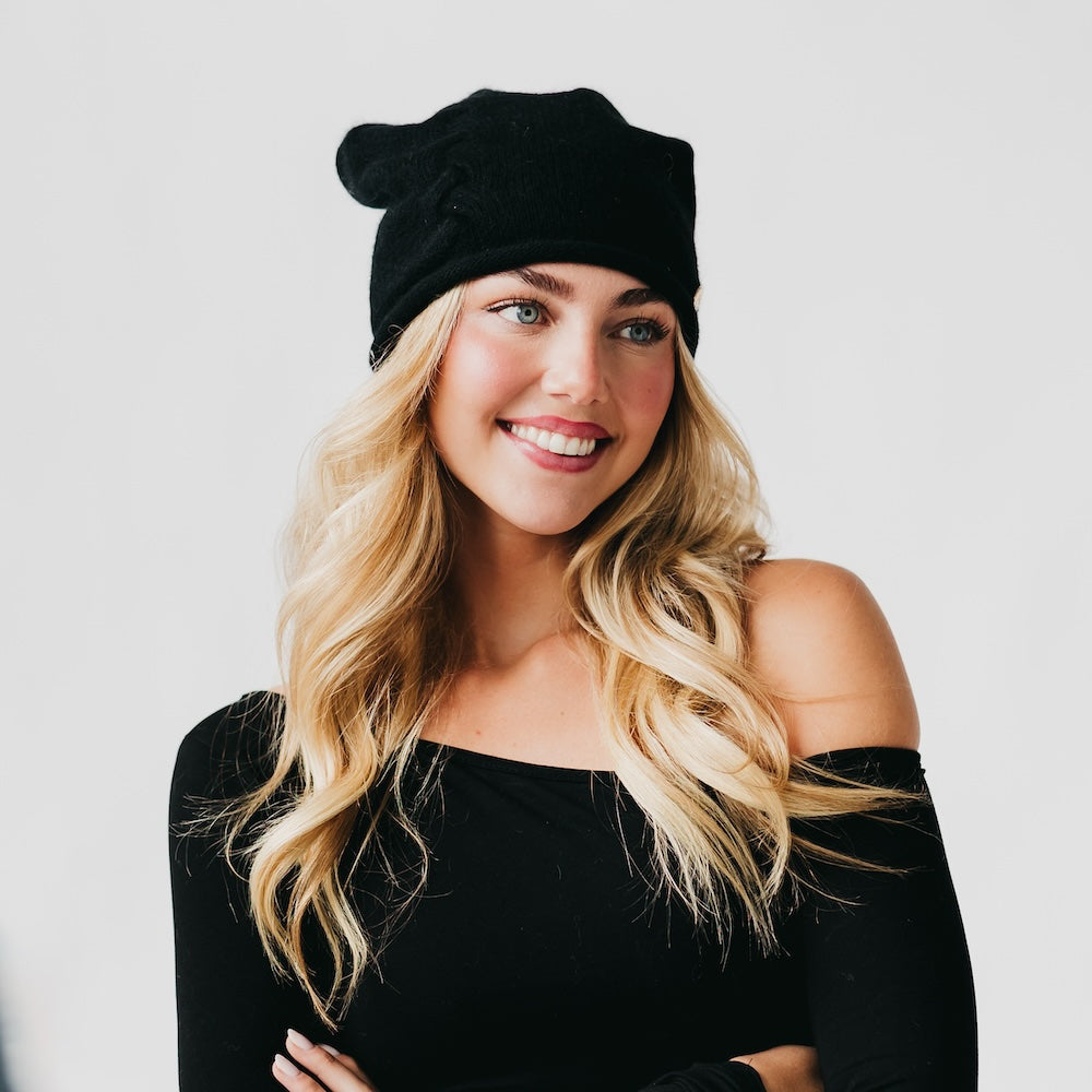 Black cashmere beanie with rolled edge and small front pin, worn on head.