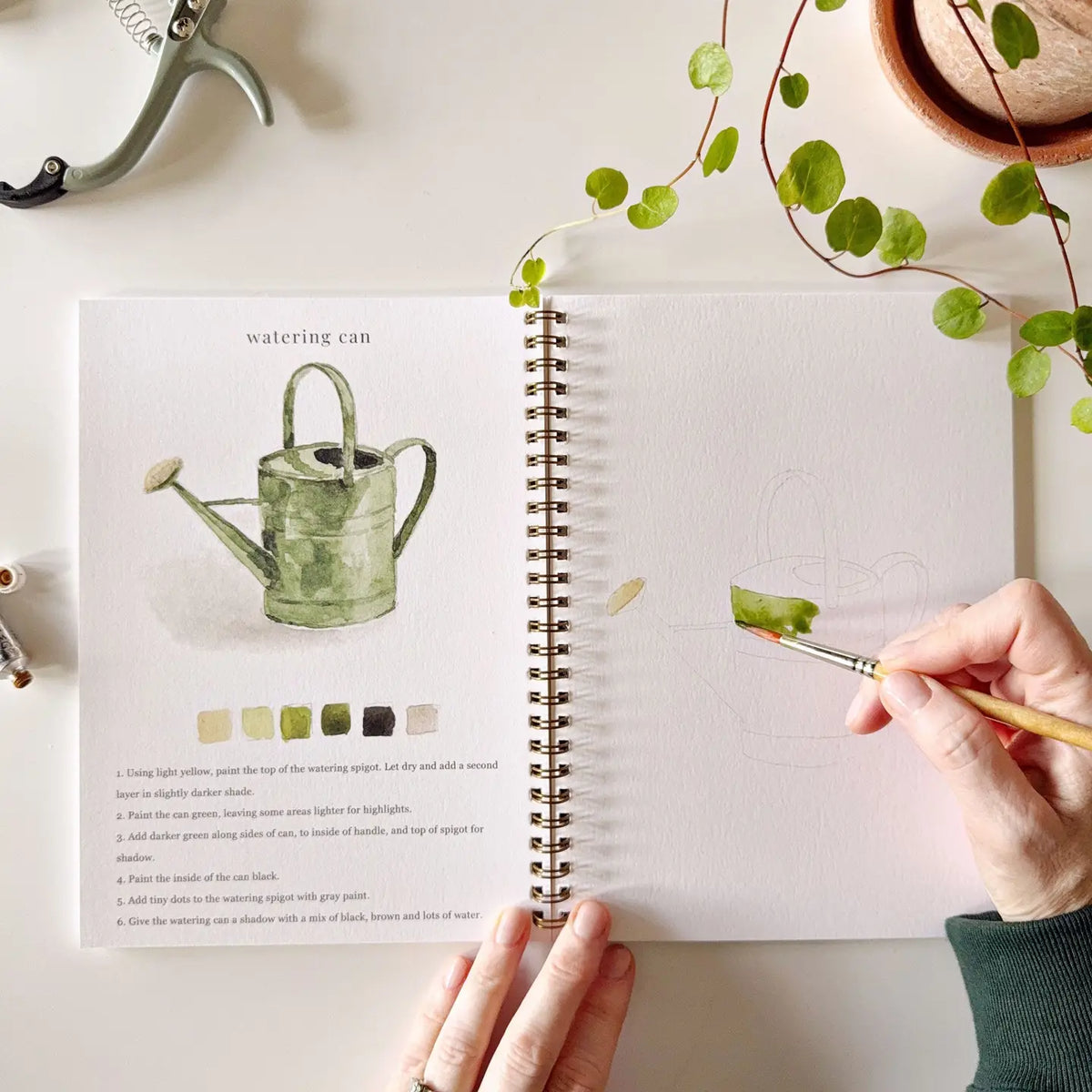Garden watercolor workbook open with watering can illustration and faint watering can outline