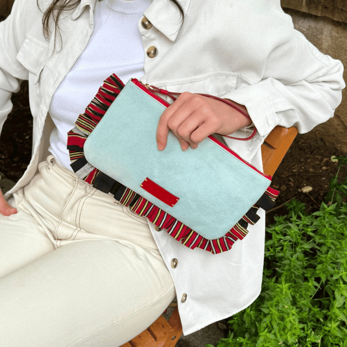 Light blue denim clutch with red zipper pull, front logo patch, and striped trim