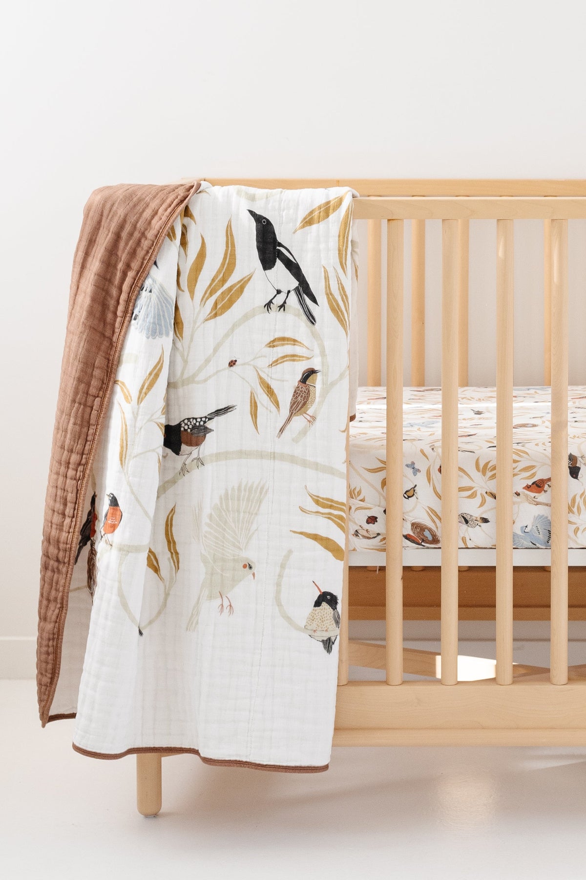 Bird-print quilt draped on crib rail, white with colorful birds and tan backing