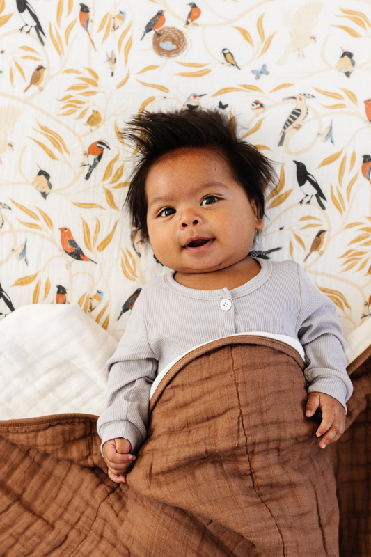 Bird-themed fitted crib sheet draped beside a baby, colorful birds and foliage print