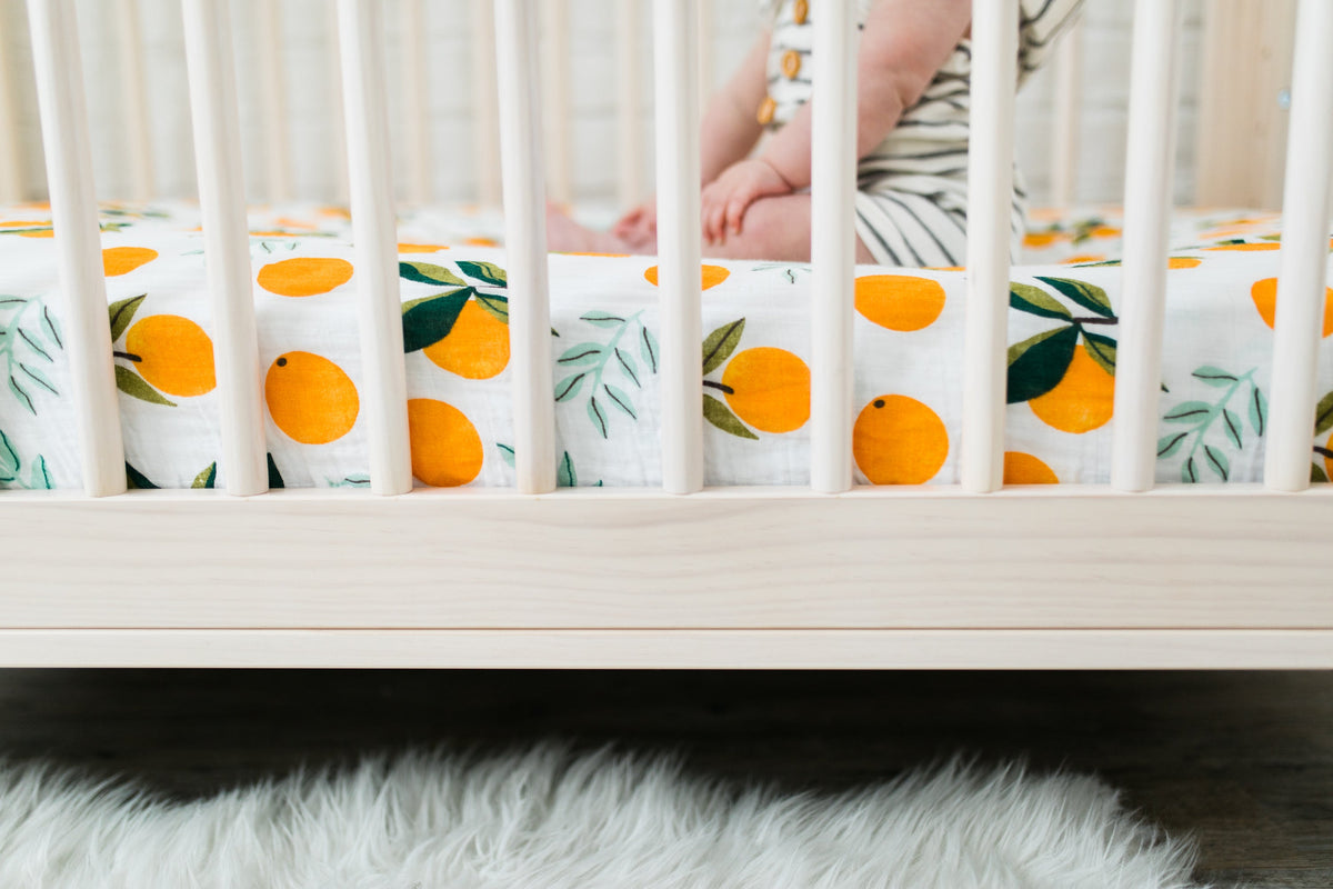 Orange citrus print crib sheet with green leaves, laid on a light wood crib