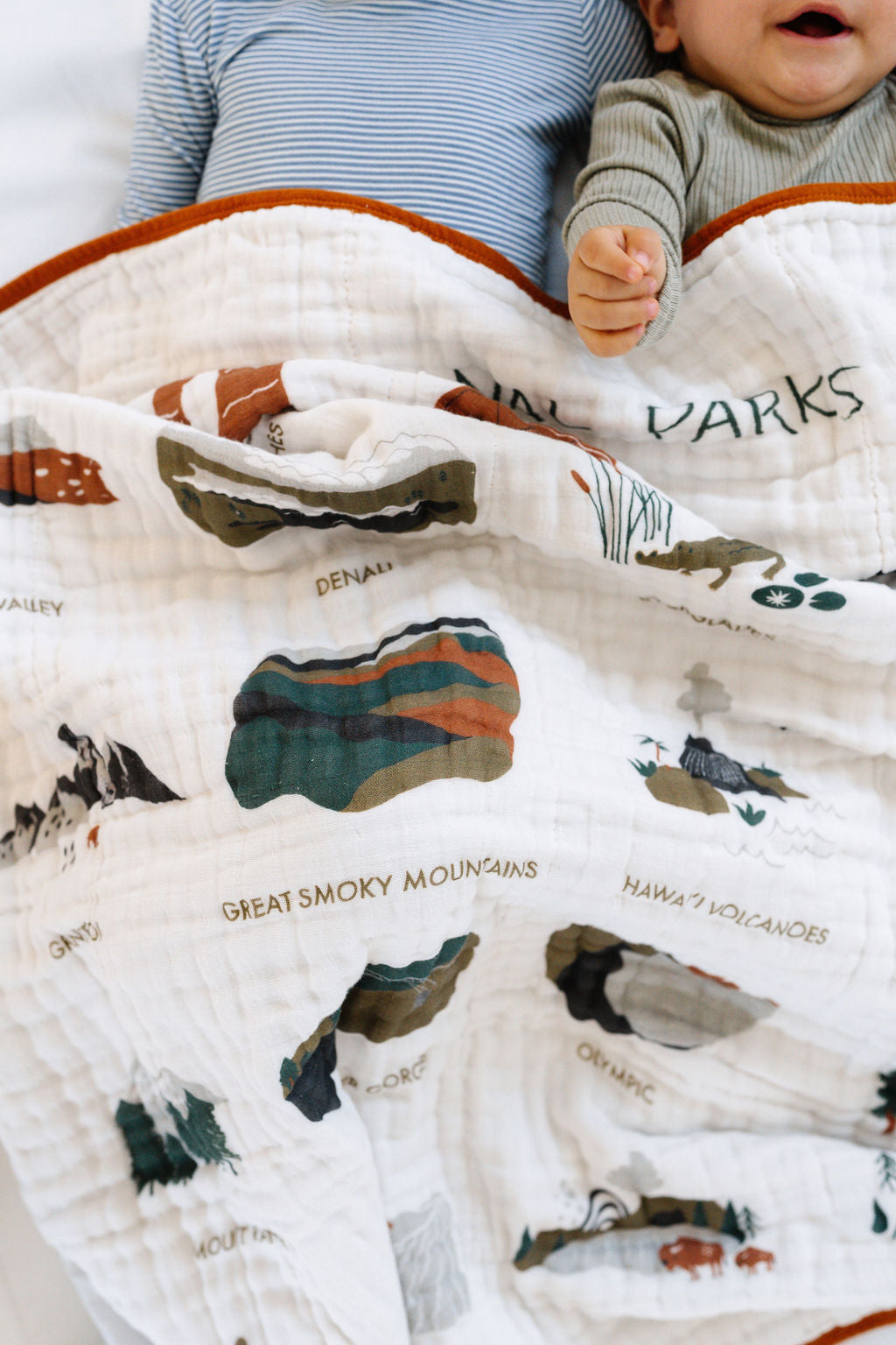 Baby holding white muslin blanket with orange trim and illustrated park names and views